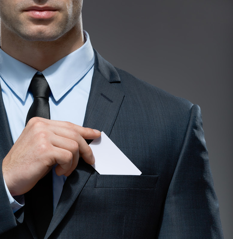 An individual wearing a suit and tie, pulling a white business card out of the suit jacket breast pocket.